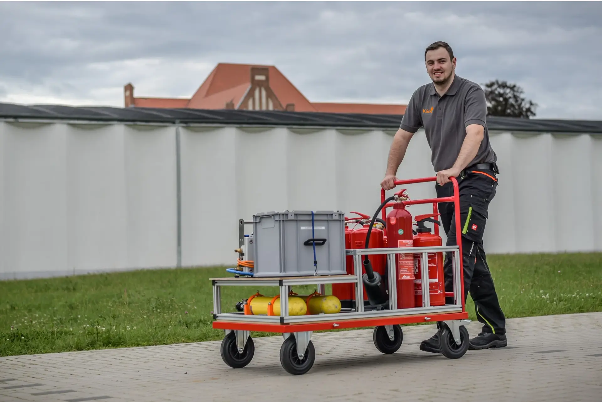 Ein Mitarbeiter schiebt einen Wagen mit Feuerlöschern, Gasflaschen und einer Transportkiste über einen gepflasterten Weg, dahinter Rasen und ein langes Gebäude