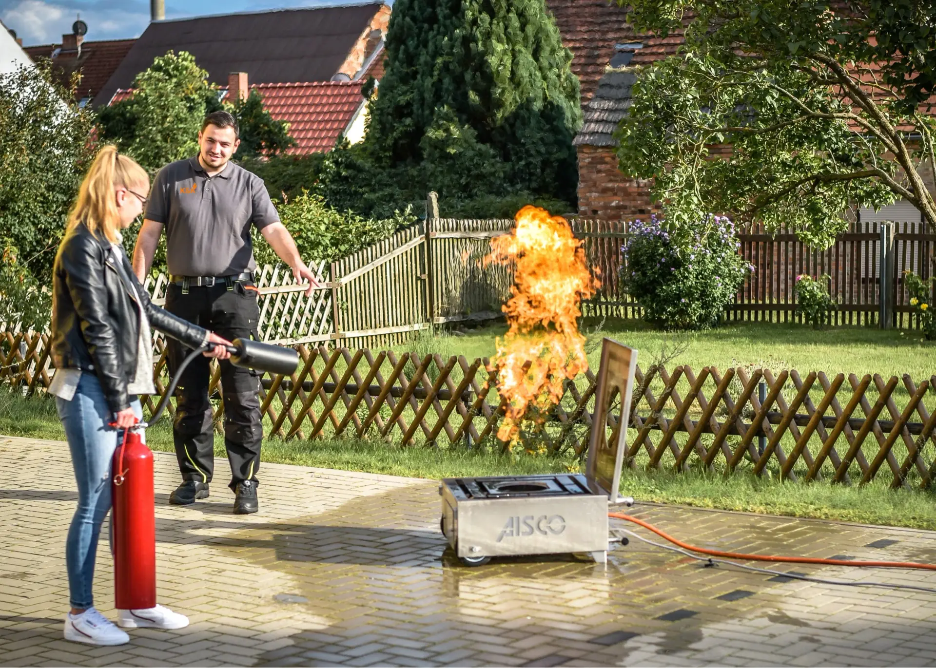 Eine Teilnehmerin zielt mit einem roten Feuerlöscher auf die Gasflamme eines Übungsgeräts aus Edelstahl, ein Ausbilder steht neben ihr und zeigt auf das Feuer